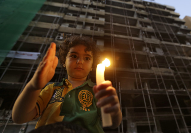 طفل لبناني يحمل شمعة أمام مبنى مدمرّ. Photo Credit: AP