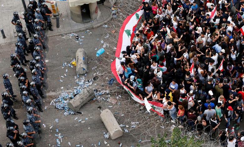 متظاهرون يرفعون الأعلام اللبنانية بمواجهة شرطة مكافحة الشغب خلف الأسلاك الشائكة خلال مظاهرة 17 تشرين الأول 2019. الصورة : محمد أزاكير/ رويترز.